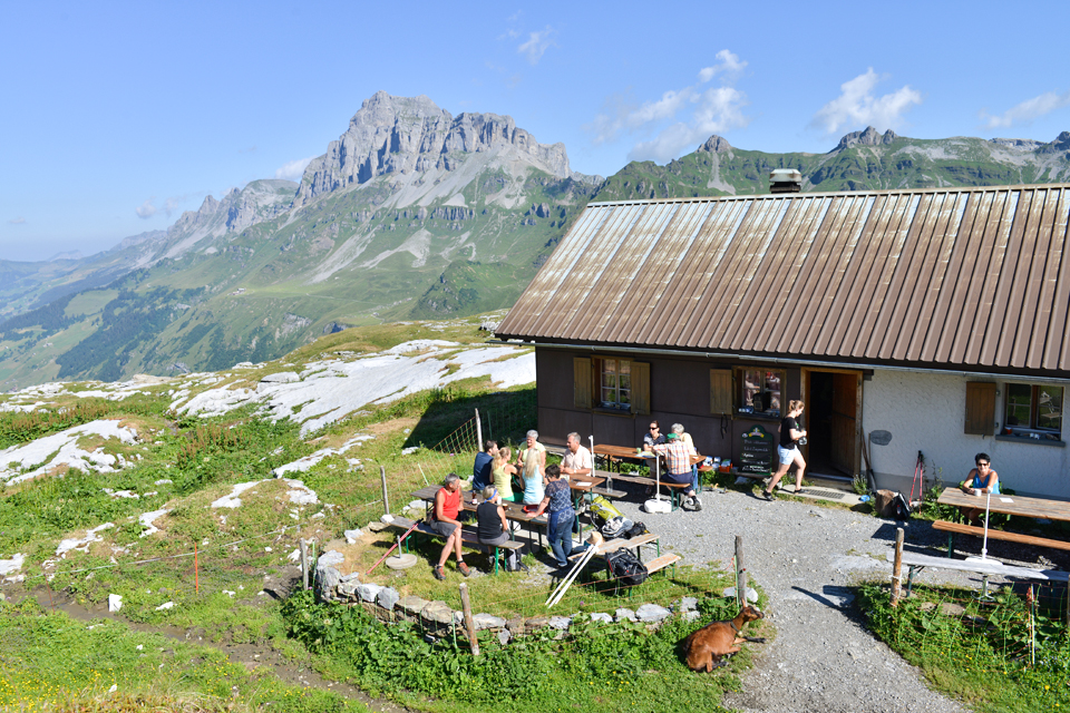 Guests sit in front of an alpine hut in the middle of a mountain panorama